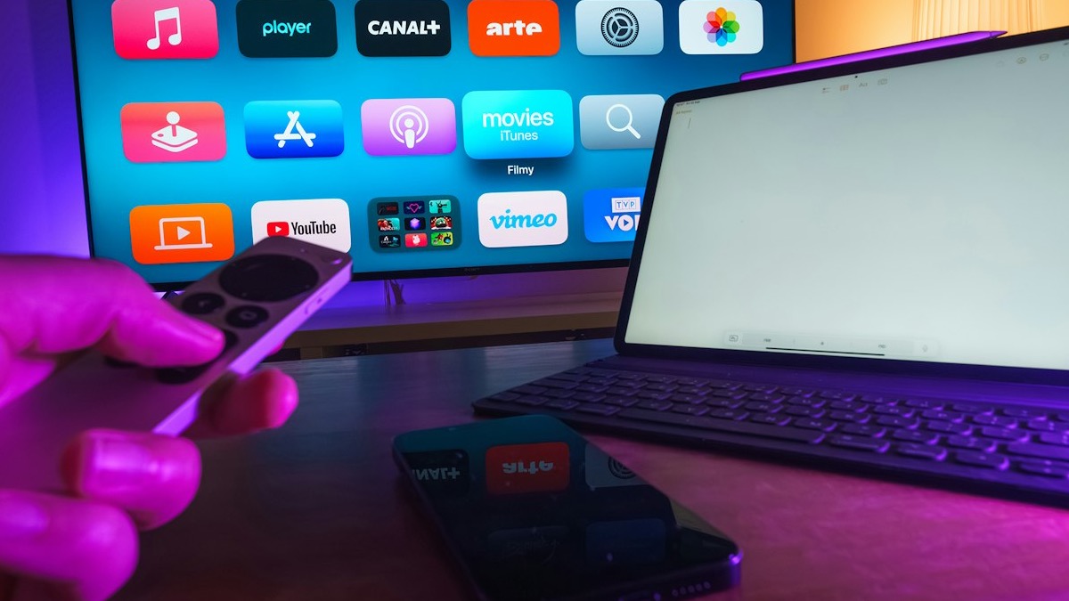 A hand holding a remote control in front of a television showing a row of app icons, illustrating how to choose a player app for FireDroid TV.